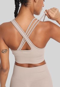 Beżowy stanik sportowy z krzyżującymi się ramiączkami i detalem w kształcie dziurki z tyłu. Teksturowana tkanina i dopasowany krój, przedstawiony z boku.