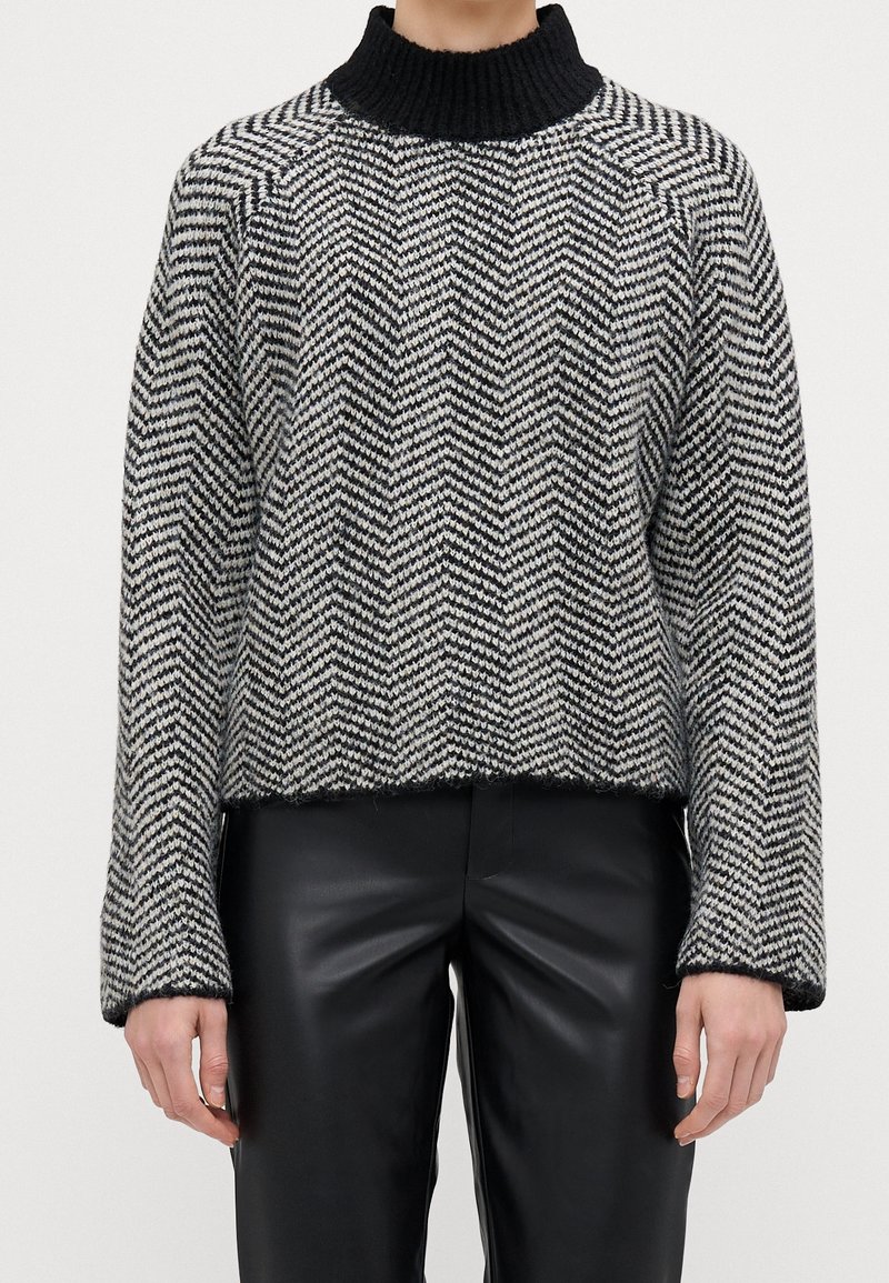 Hřejivý pletený svetr s černým stojáčkem, zdobený šedobílým herringbone vzorem. Hladké manžety a spodní lem, jemně texturovaný.