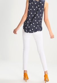 Marineblauw mouwloos topje met witte stippen, gecombineerd met witte slim jeans en oranje hoge hakken. Glad materiaal en getailleerd ontwerp.