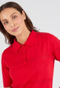 Rode gebreide polo shirt met een geschulpte kraag, korte mouwen en een textuurpatroon door de stof. Detail van vier knopen.
