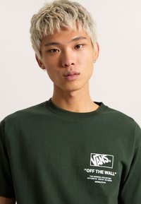 Camiseta de algodón verde con un logo blanco de "VANS" y el texto "OFF THE WALL" en el pecho. Manga corta, cuello redondo, corte estándar.
