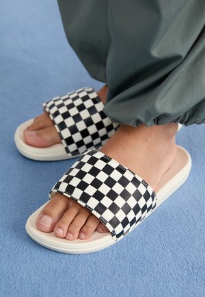 Sort og hvidt ternet mønster på sliders med flad beige sål, der har en bred rem og tekstureret materiale.