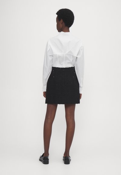 Chemise blanche à manches longues et jupe noire texturée, présentant une silhouette ajustée et des coutures visibles, portée avec des chaussures noires.