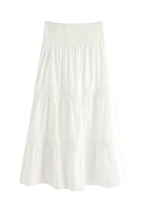 Nuori nainen, jolla on vaaleanvioletti t-paita, valkoinen kerroksellinen maksihame, mustat sandaalit, valkoinen röyhelöinen käsilaukku kädessä, hymyilevä ja seisoo valkoisella taustalla.