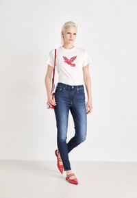 Cremefarbenes T-Shirt mit rotem Vogel-Graphic, kombiniert mit blauen Skinny Jeans und roten spitzen Ballerinas. Das Model hält eine kleine rote Umhängetasche.
