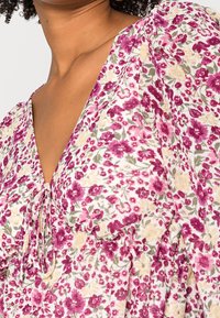 Gros plan d'une personne portant une robe fleurie avec des fleurs roses, violettes et beiges, dotée d'un col en V et d'un détail froncé au niveau de la poitrine.