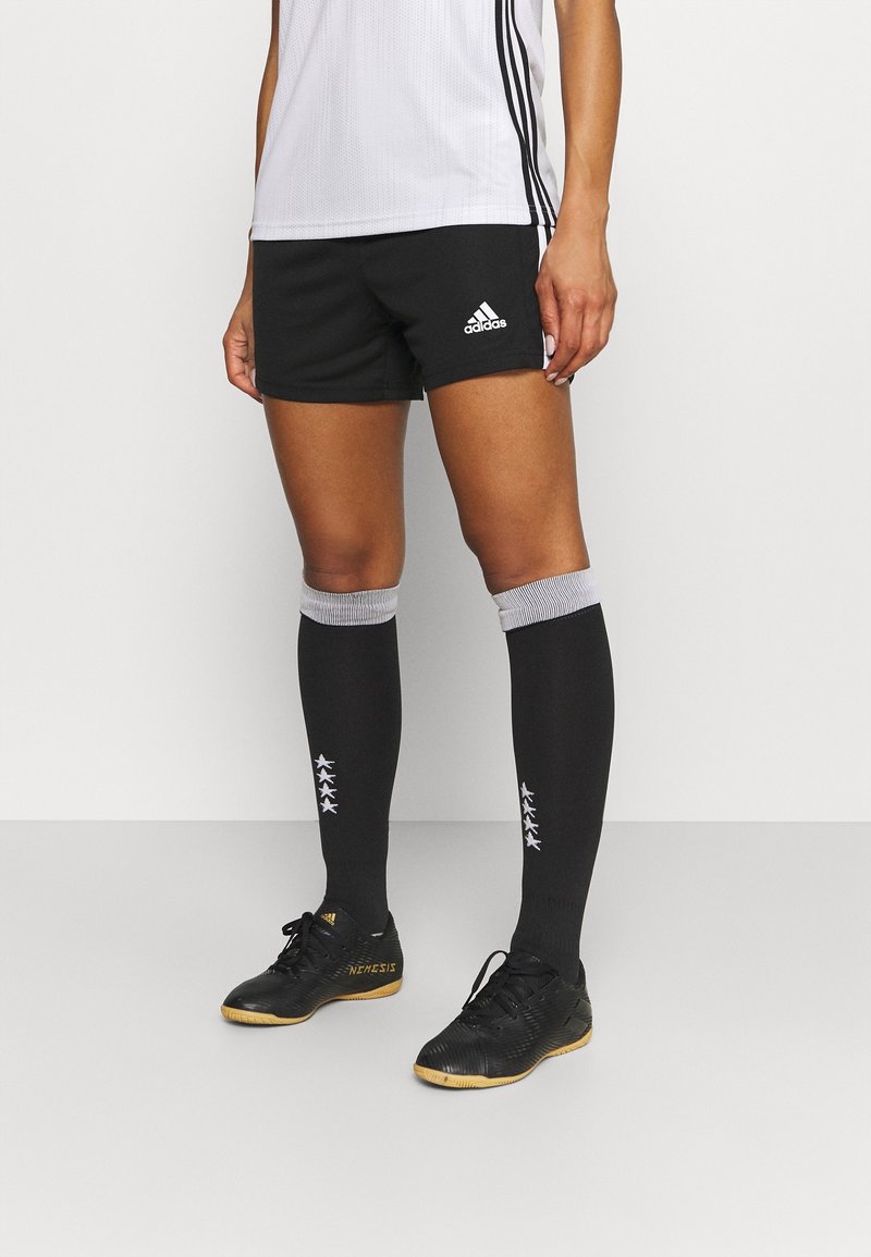 Persona que lleva pantalones cortos negros de Adidas, calcetines negros hasta la rodilla con estrellas blancas, zapatos de fútbol indoor negros y una camiseta deportiva blanca.