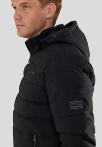 Chaqueta negra acolchada con capucha, con textura acolchada, bolsillos con cremallera y un parche de marca en la manga.