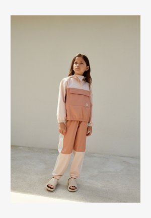 Lekka kurtka w blokowych kolorach w pastelowym różu i brzoskwiniowym odcieniu z przednią kieszenią, zestawiona z pasującymi spodniami i beżowymi klapkami.