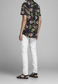 Camicia a maniche corte a fiori nera con motivi rossi e verdi, abbinata a pantaloni bianchi e sandali neri, vista da dietro.