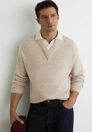 Mężczyzna w beżowym swetrze z dekoltem w serek na białej koszuli z kołnierzykiem, ciemnych dżinsach, trzyma złożony bordo sweter, stoi na tle białej ściany.