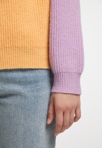 Maglione a maglia con texture a costine, caratterizzato da un corpo giallo e maniche lavanda. Abbinato a jeans di denim blu chiaro.