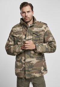 Maskovacia bunda s vysokým golierom, dlhými rukávmi a viacerými prednými vreckami. Vyrobená z látky s kombináciou zelených a hnedých odtieňov.