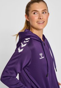 Felpa zip con cappuccio viola, dotata di cappuccio con cordino, caratterizzata da strisce bianche a zigzag sulle spalle e un logo sul petto. Tessuto morbido.