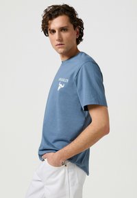 Camiseta de algodón azul para hombre con manga corta, que presenta el texto "WRANGLER" en blanco y un gráfico de un toro en el delantero. Diseño sencillo, ajuste relajado.