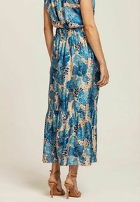 Maxiklänning i ljusbeige med stora blå blommönster, rynkad midja, volangkant och bärs med tan-färgade högklackade sandaler.