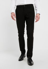 Man som bär svarta slim-fit byxor, svarta dress skor och en vit långärmad skjorta med knappar framför en enfärgad bakgrund.