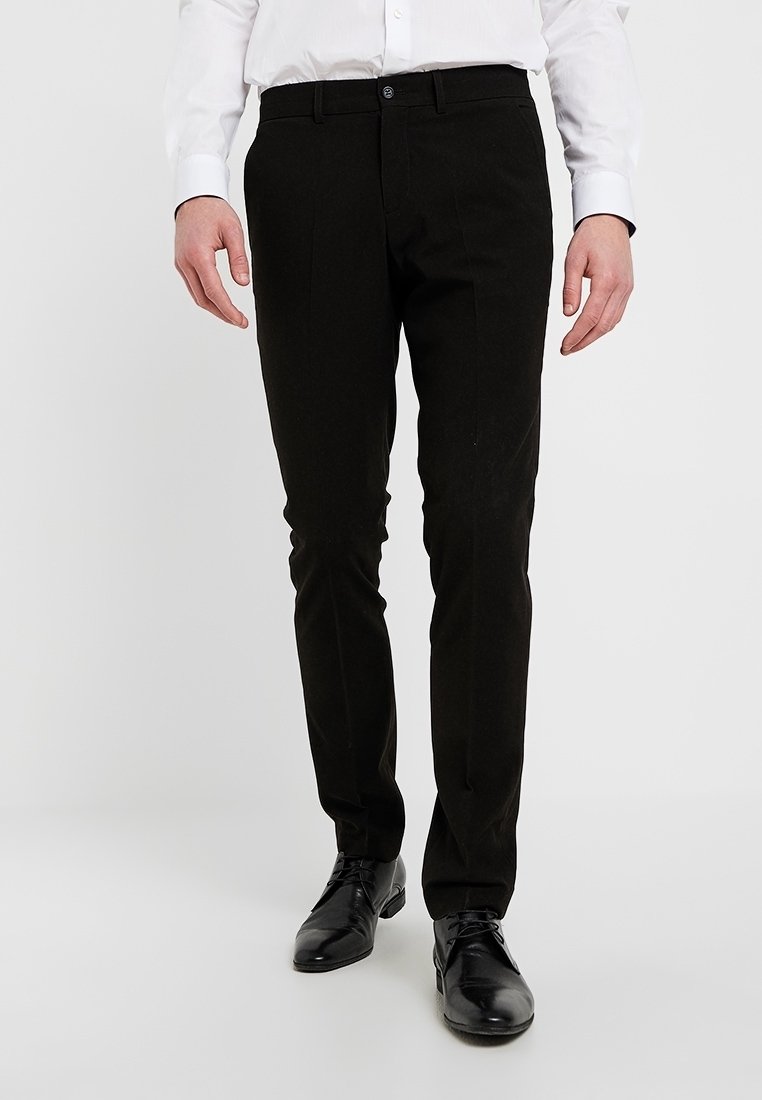 Man som bär svarta slim-fit byxor, svarta dress skor och en vit långärmad skjorta med knappar framför en enfärgad bakgrund.