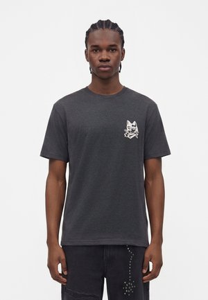 Jeune homme aux cheveux tressés portant un t-shirt gris foncé avec un petit dessin de chat blanc sur la poitrine gauche.