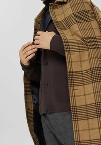 Braun kariertem Oberbekleidung mit strukturierter Oberfläche, drapiert über einem dunkelgrauen Cardigan mit Knöpfen und einer Taschendetail.