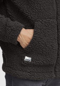 Giacca in fleece nera con una superficie testurizzata e soffice, dotata di una tasca laterale e di un'etichetta che mostra la scritta "11 PROJECT."