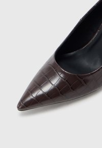 Καφέ γόβα με μοτίβο κροκοδείλου, μυτερής μύτης, με λεία υφή και μαύρο εσωτερικό. Κομψός σχεδιασμός που εστιάζει στη λεπτή γραμμή και το υλικό.