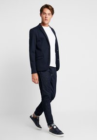 Blazer navy de dos botones sobre una camiseta blanca, combinado con jeans oscuros y zapatillas navy, con puños doblados y tejido texturizado.