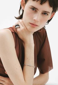 Anillo textural en negro y gris, pulsera de plata con piedras transparentes, llevada por una persona con cabello corto y top marrón, sobre un fondo blanco.