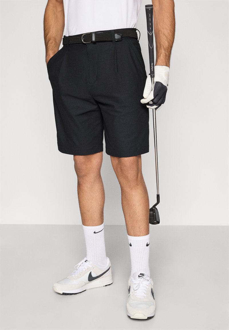 Shorts de golf noirs avec une ceinture tressée, associés à des chaussettes blanches et des baskets. Un club de golf est tenu dans une main, avec un gant à l'autre.