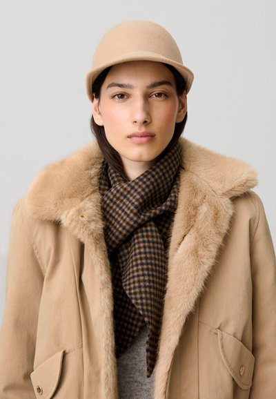 Mujer con sombrero beige, abrigo marrón con cuello de piel y bufanda oscura a cuadros, mirando hacia adelante con expresión neutral sobre un fondo liso.