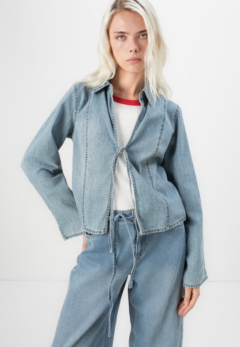 Lichtblauwe spijkerjacket met striksluiting, lange mouwen en gestikte details. Draagt over een witte top met rode accenten en gecombineerd met bijpassende broek.