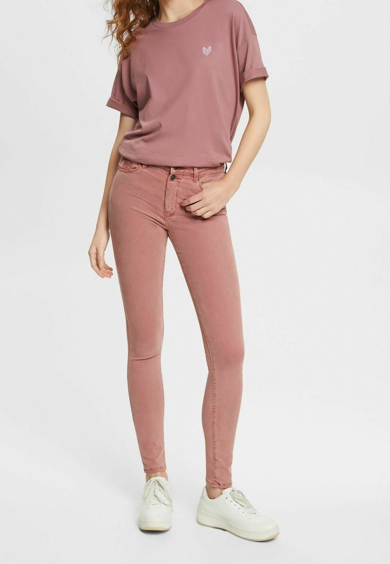 Femme portant un jean moulant rose poudré, un t-shirt ample assorti avec un petit emblème en forme de cœur, et des baskets blanches sur un fond uni.
