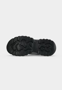 Semelle de chaussure en caoutchouc noir avec des motifs de bande de roulement profonds et irréguliers conçus pour l'adhérence et la durabilité sur fond gris clair.