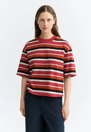 Jonge vrouw met lang bruin haar, gekleed in een rood, zwart en wit gestreept T-shirt met korte mouwen en een donkere broek, staand tegen een effen achtergrond.