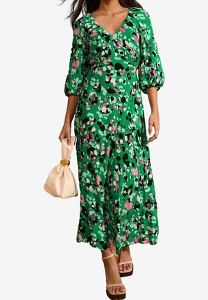 Donna che indossa un abito midi verde a fiori con maniche a sbuffo, tiene in mano una borsa da sera beige e indossa sandali con plateau beige e cinturini.