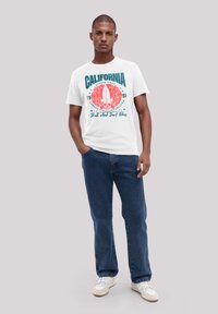 White t-shirt featuring a colorful "California" graphic with palm trees, surfboard, and skull motifs. Worn with dark blue jeans and white sneakers.