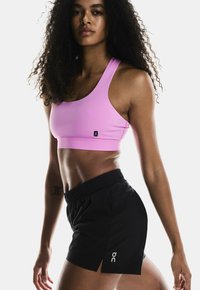Sujetador deportivo rosa y pantalones cortos negros con aberturas laterales; la tela parece suave; logo minimalista en los pantalones cortos; la modelo tiene el cabello rizado.