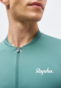 Κοντινό πλάνο στον λαιμό και το κάτω μέρος του προσώπου ενός άνδρα που φοράει μια τιρκουάζ ποδηλατική φανέλα Rapha με φερμουάρ μπροστά και μικρό κρίκο στο αυτί.