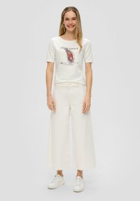 T-shirt blanche avec un imprimé paisley coloré et du texte, associée à un pantalon large blanc et des baskets blanches, sur un fond neutre.