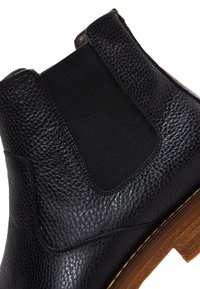 Bottine Chelsea en cuir noir avec une tige texturée, des panneaux latéraux élastiques et une semelle en bois. Présente un bout arrondi et un détail de couture subtil.