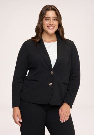 Donna sorridente con lunghi capelli castani indossa un blazer nero con bottoni dorati sopra una maglietta bianca, in piedi davanti a uno sfondo chiaro e semplice.