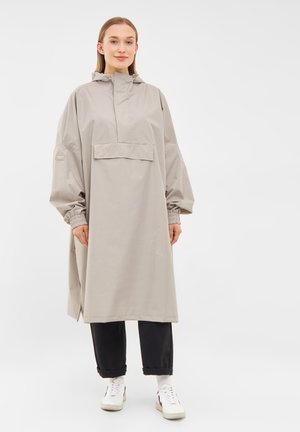 Anorak impermeabil bej deschis, cu glugă, buzunar frontal, manșete elastice și croi lejer. Asortat cu pantaloni închiși la culoare și adidași albi.
