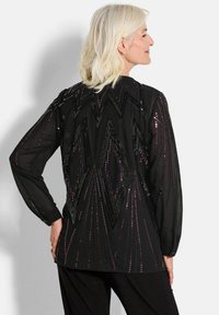 Schwarze Bluse mit geometrischem Design und Pailletten in verschiedenen Lila-Tönen. Lange Ärmel und eine lockere Silhouette. Glatte Stoffstruktur.