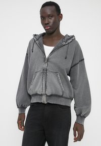 Hanorac gri cu fermoar, realizat dintr-un material moale. Include glugă, buzunar tip cangur și manșete elastice. Marginile netede și cusăturile contrastante adaugă detalii.