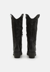 Par de botas de vaquero de cuero negro hasta la pantorrilla con tiradores y tacones cuadrados, mostradas desde atrás sobre un fondo blanco liso.