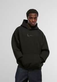 Musthaveed kapuutsiga fliisist dressipluus, millel on ees suur känguru tasku ja valge allkirjalogo rinnal. Sile tekstuur.