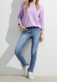 Haut long à manches longues texturé de couleur violet clair associé à un jean ajusté bleu moyen. Le mannequin porte des baskets blanches avec des accents rouges. Tenue simple et décontractée.