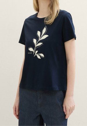 Kobieta nosząca granatowy t-shirt z krótkim rękawem z beżowym wzorem liścia i logo "Tom Tailor", w połączeniu z ciemnoniebieskimi spodniami.