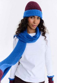 Zestaw czapka i szalik z niebieskiej i bordowej wełny; czapka ma mankiet i zielony akcent. Szalik jest długi i gładki, ułożony na białej koszuli.