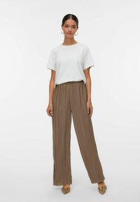 Witte T-shirt met korte mouwen, gecombineerd met bruine geplooide wijde broek en gepatternde puntige schoenen, staand tegen een lichte achtergrond.